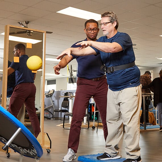 A man is standing next to his therapist and throwing a ball onto a trampoline to practice coordination