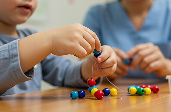 A close up of a young boy's hands as he is placing beads on a string with an adult in the background